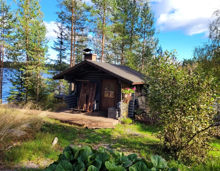 Small wooden cabin surrounded by trees near a lake on a sunny day.