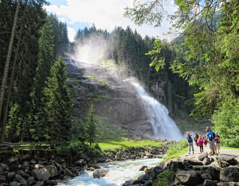 People hiking near a waterfall surrounded by tall trees and rocky stream.