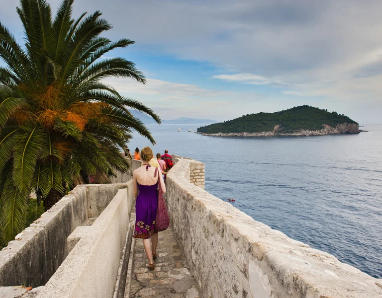 Woman walking along a stone pathway by the sea with a palm tree and island in the distance.