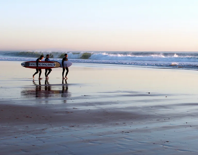 Three surfers carrying boards along a wet sandy beach at sunset.