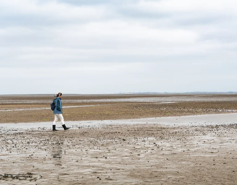 Person walking alone on a vast, cloudy beach at low tide.