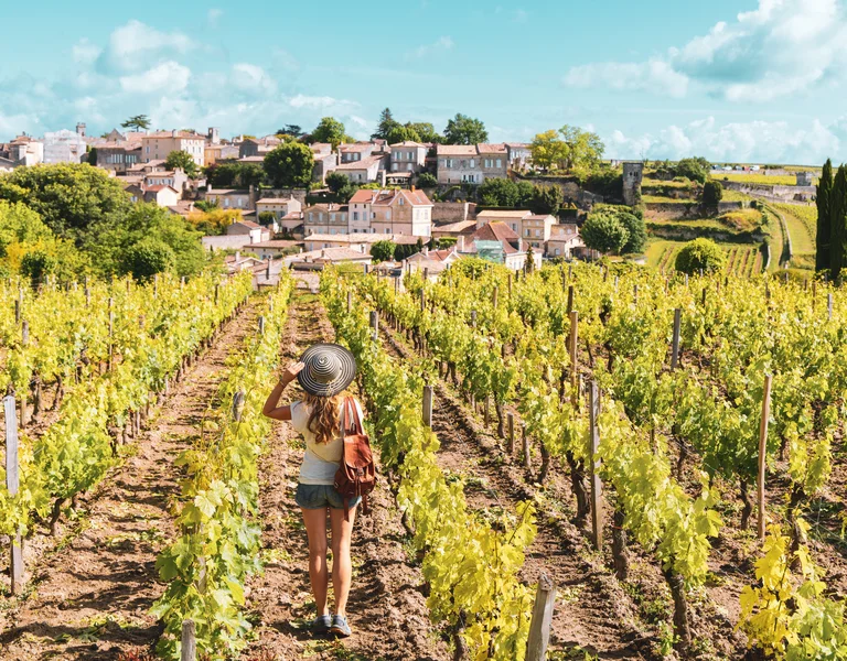 Woman walking through a vineyard with a village in the background under a blue sky.