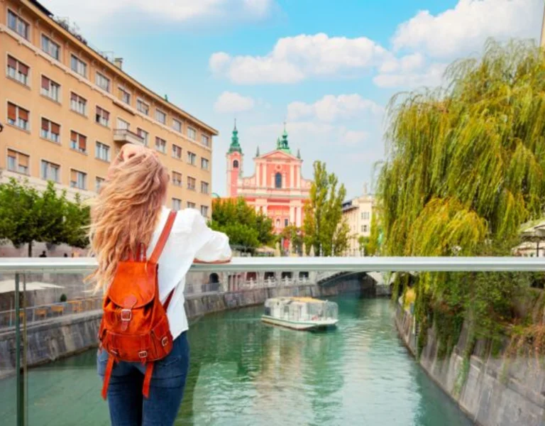 Woman with a red backpack stands on a bridge overlooking a river in a city.