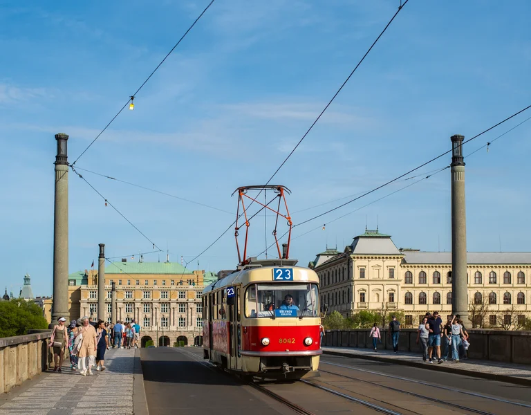Red and cream tram crossing a city bridge under a blue sky, people walking nearby.