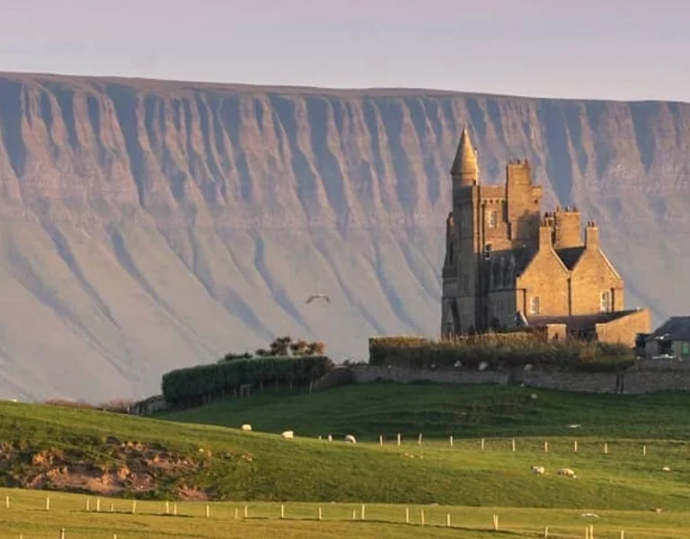 Castle-like building on green fields with sheep, set against a steep, flat-topped mountain.