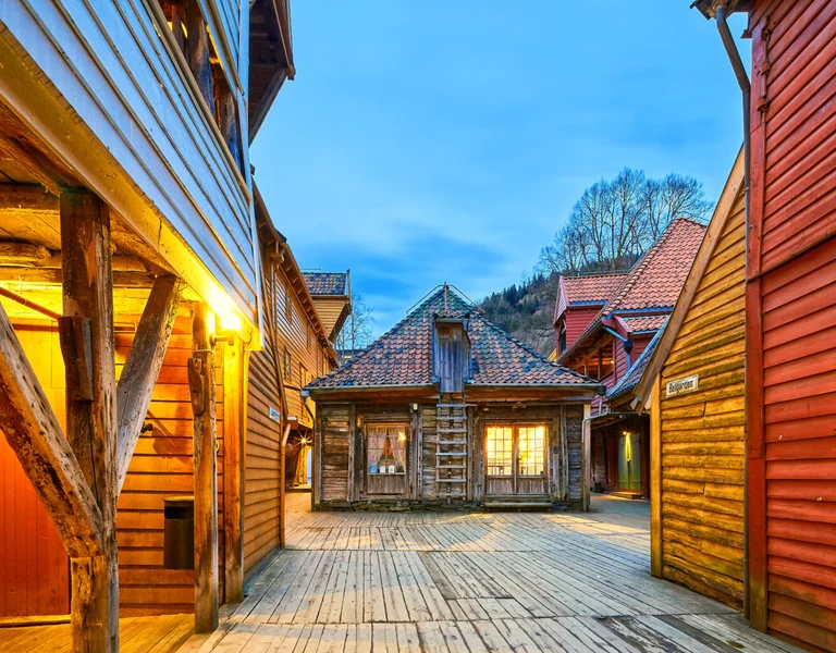 Wooden alley with colorful houses at dusk, warm lights shining from windows.