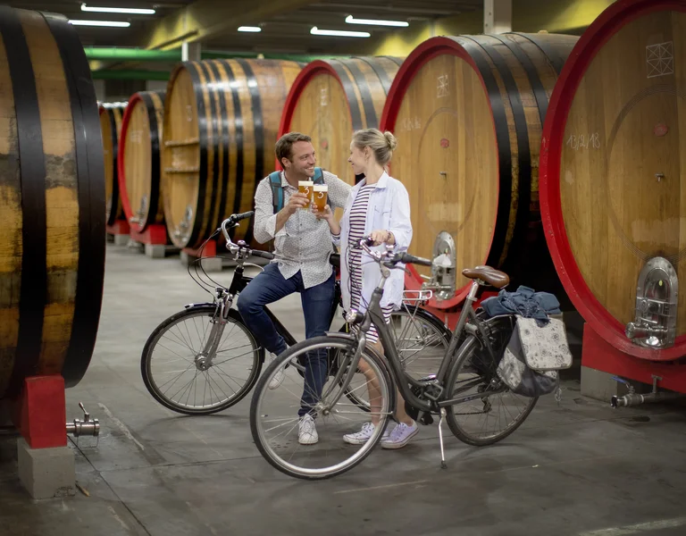 Two people with bicycles talk and smile inside a winery with large wooden barrels.