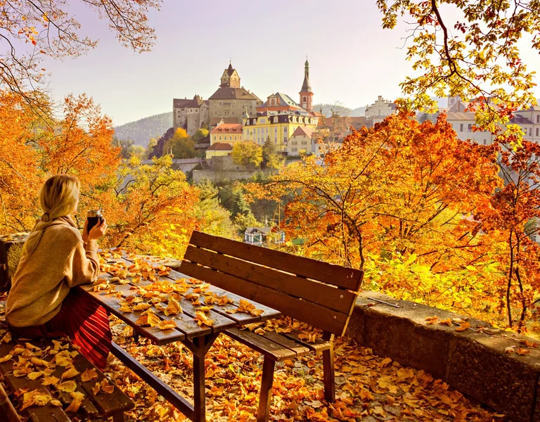 Woman sitting on a bench in a park with autumn leaves, overlooking a scenic town.