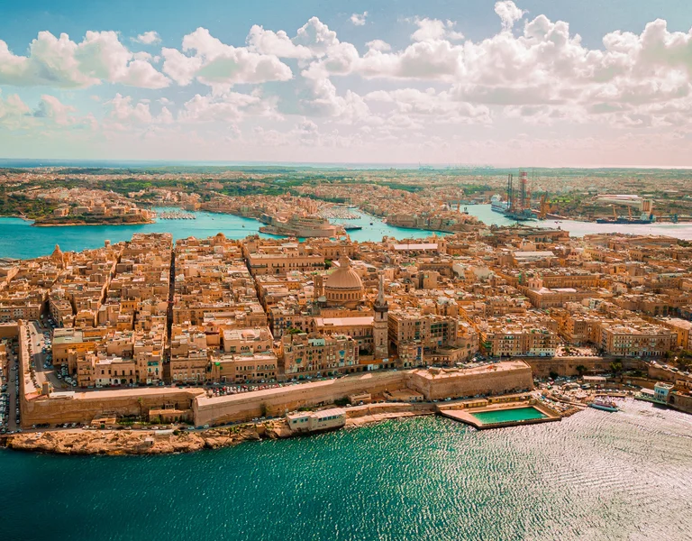 Aerial view of a coastal city with tan buildings and turquoise water under a bright sky.