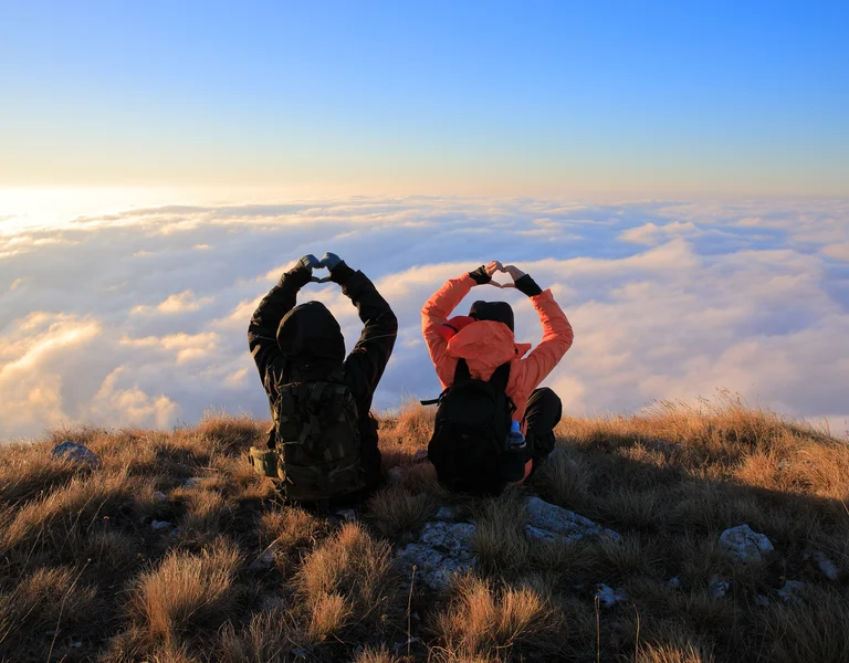 Two people sit on a grassy hilltop taking photos of clouds at sunrise.