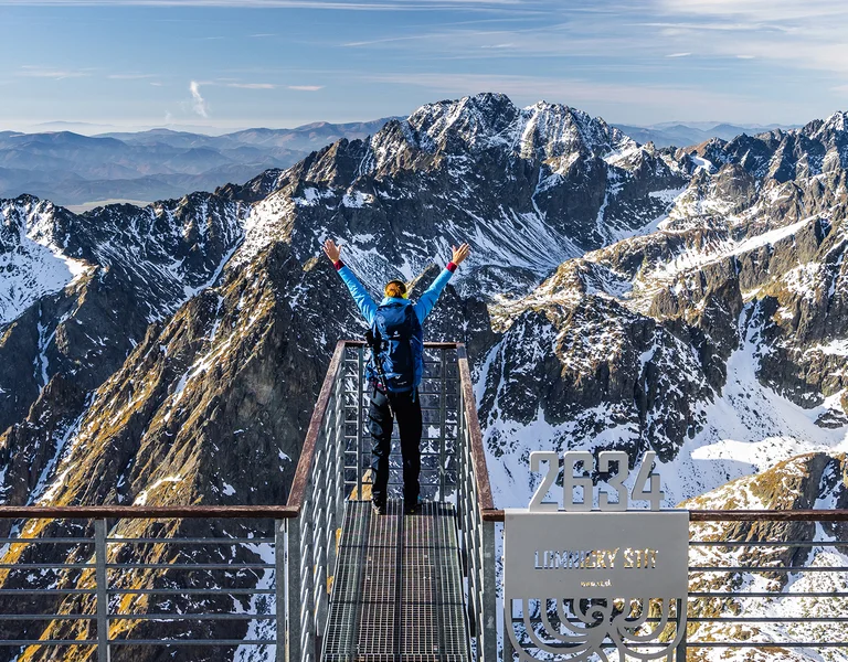 Two people stand on a viewing platform, arms raised, overlooking snow-capped mountains.