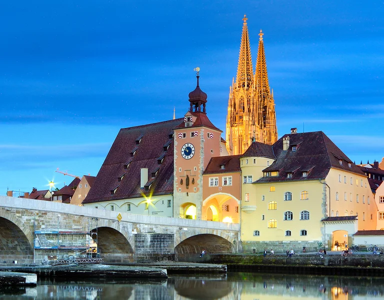 Stone bridge over a river with illuminated historic buildings and a cathedral at dusk.