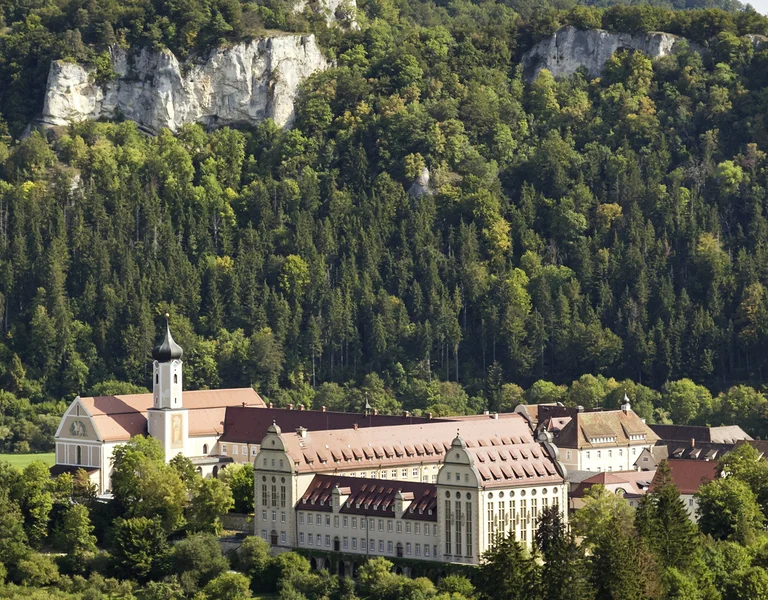 Large monastery with red roofs nestled in a green valley, surrounded by forested hills.