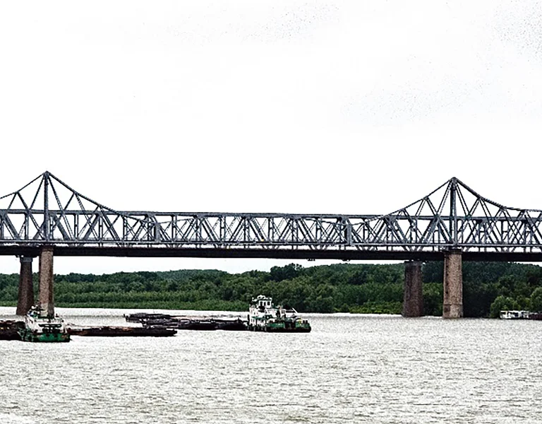 Steel truss bridge spanning a wide river with boats and trees on a cloudy day.
