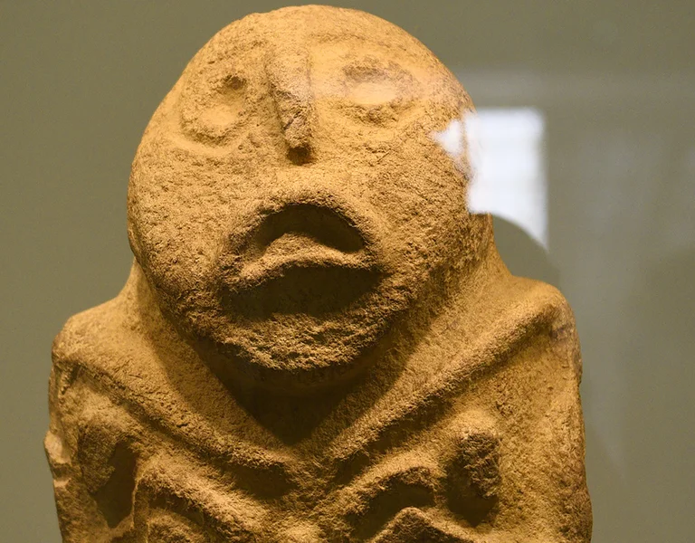 Stone sculpture of a figure with wide eyes and an open mouth, arms crossed on chest.