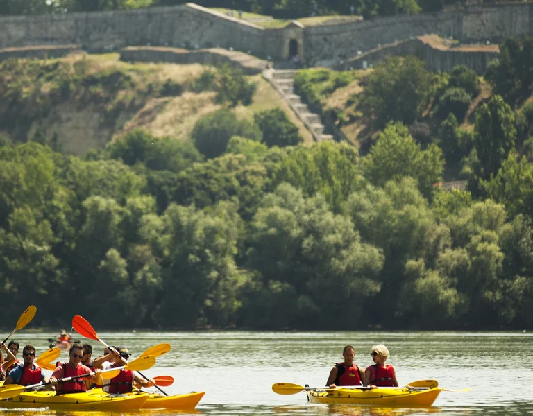 People kayaking on a calm river with a forested hillside and castle in the background.