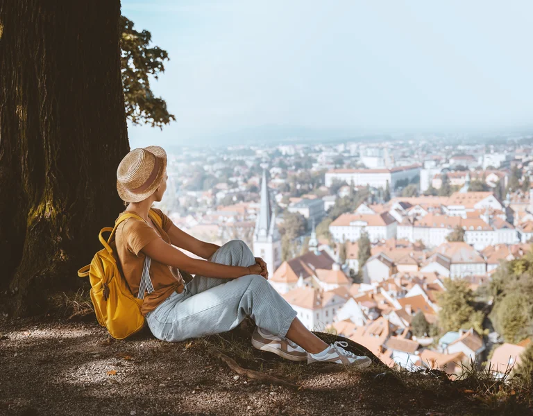 Woman with a yellow backpack sits under a tree, overlooking a city with red rooftops.