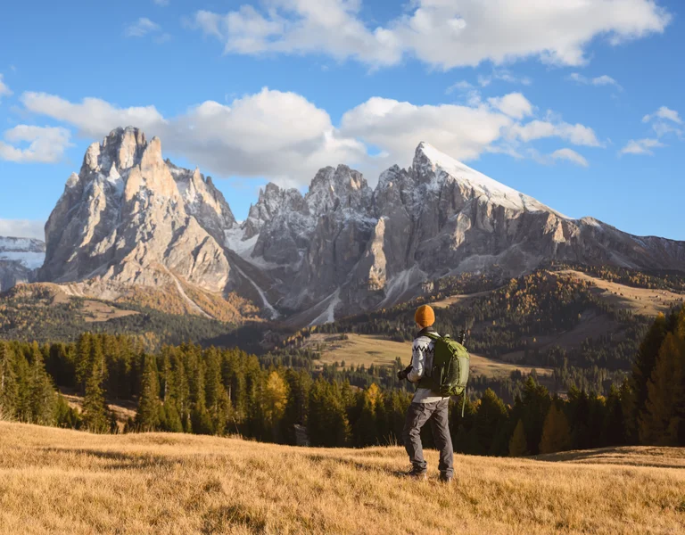 Hiker with a backpack looks at snow-capped mountains under a blue sky.