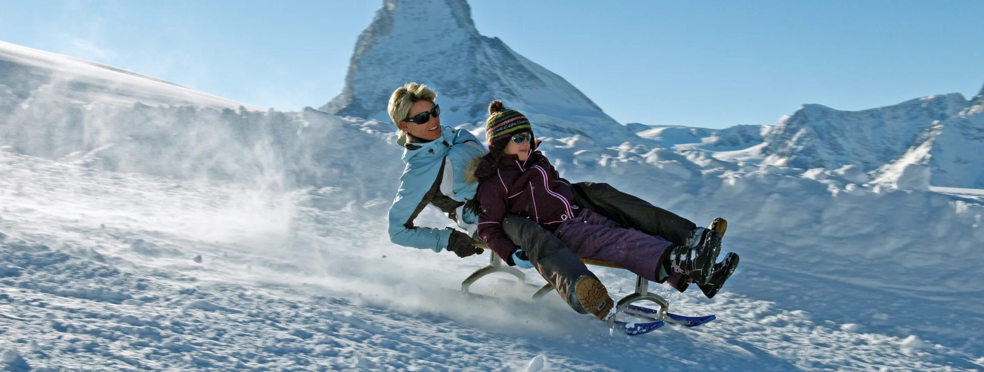 Two people sledding on snow with a mountain peak in the background.