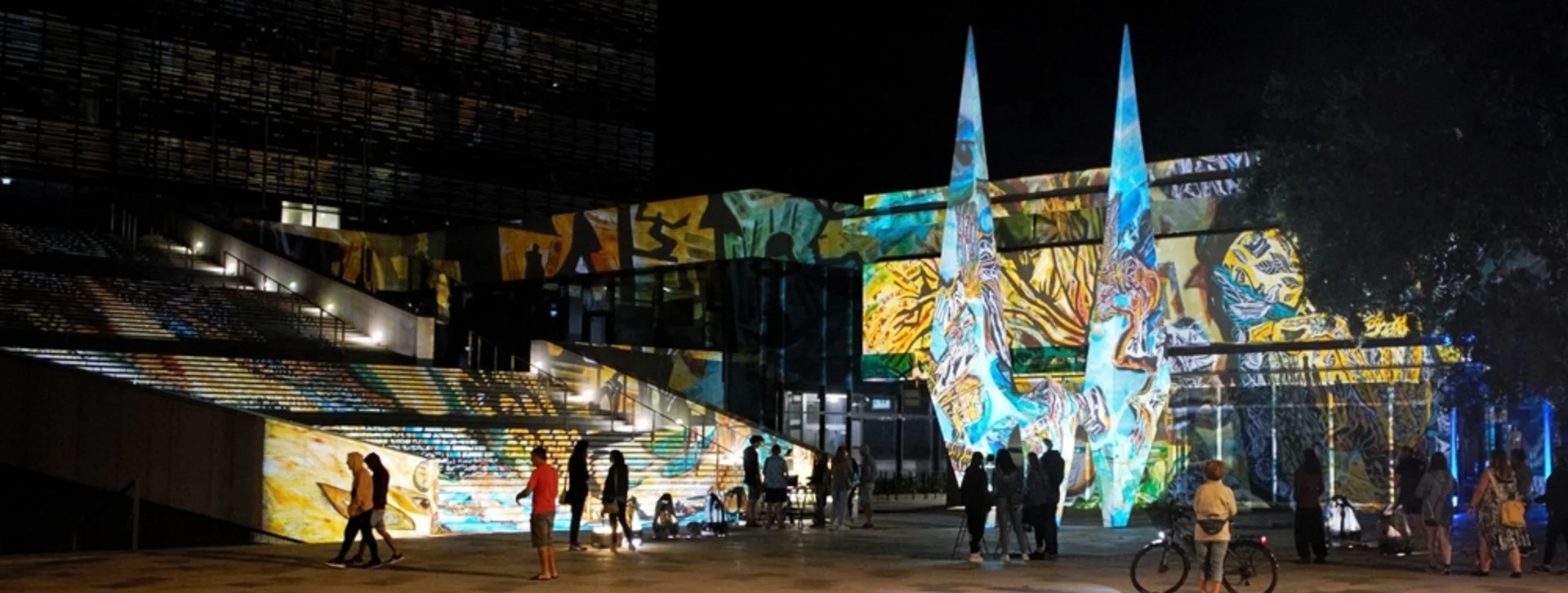 Night scene of a modern building with colorful light projections and people milling about.