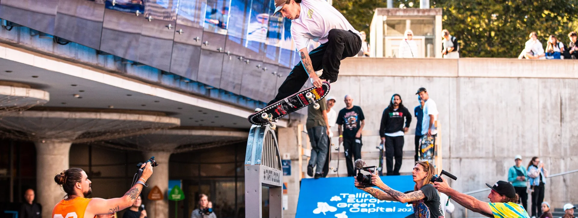 A skateboarder performing a trick on a blue ramp with "Visit Estonia" branding, surrounded by spectators and photographers at an outdoor event.