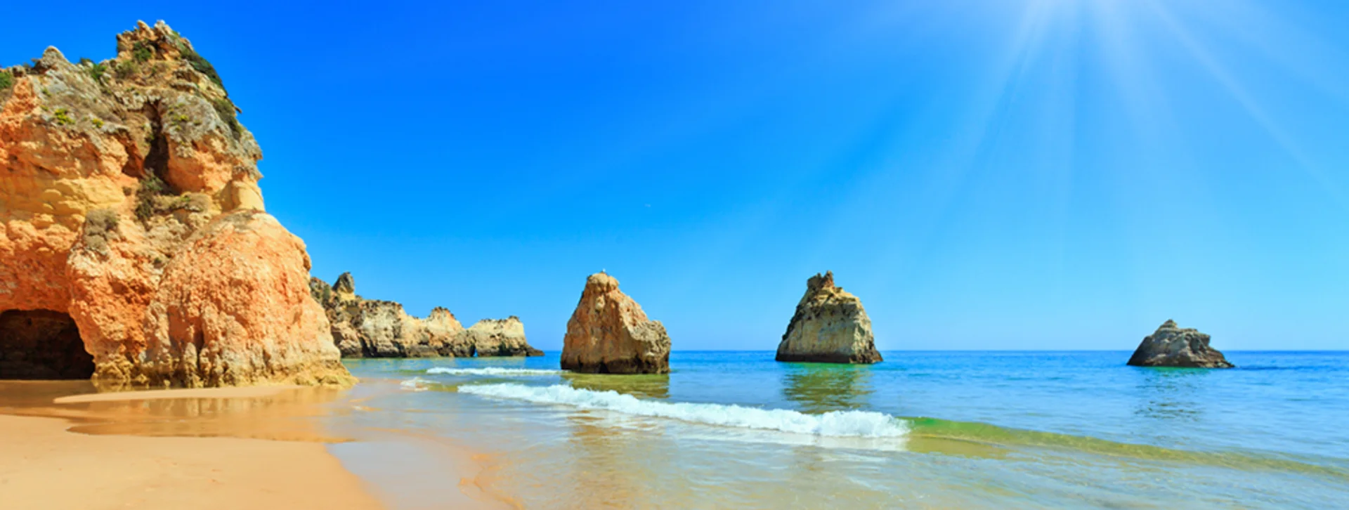 Sunny beach with golden cliffs and rock formations in clear blue sea.