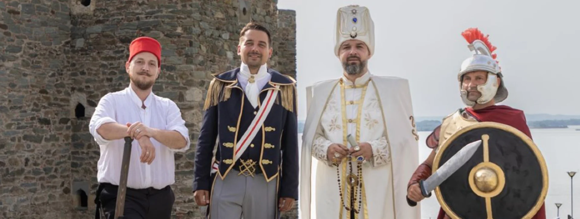 Four people in historical costumes stand in front of a stone wall.
