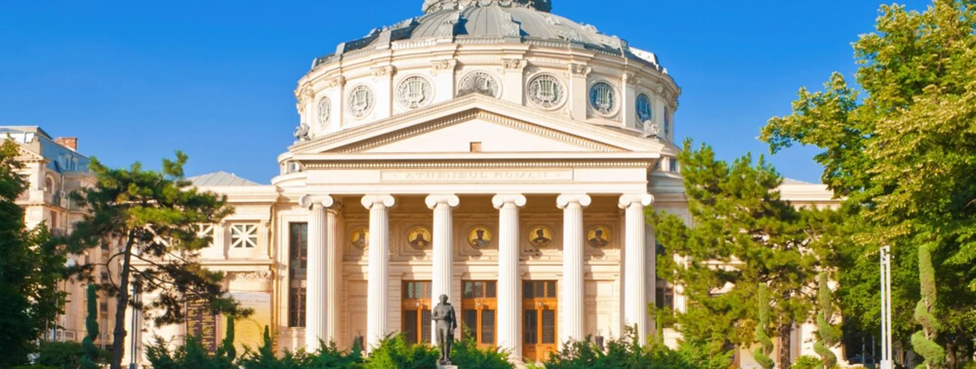 Romanian Athenaeum, a building with columns and dome, surrounded by green trees under a clear sky.