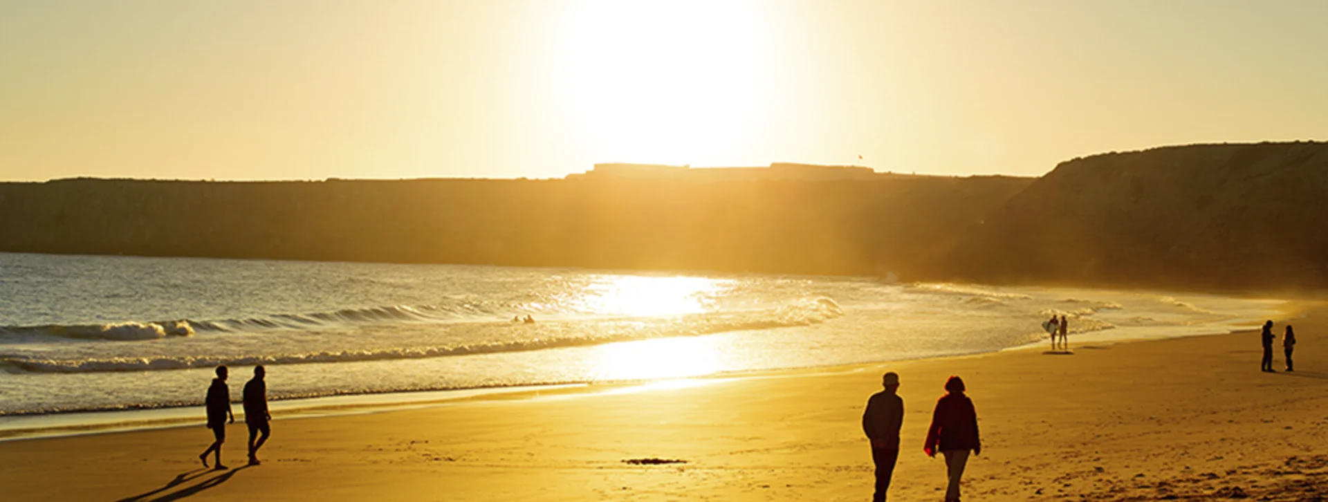 People walking on a beach in Algarve, Portugal, during sunset with cliffs in the background.