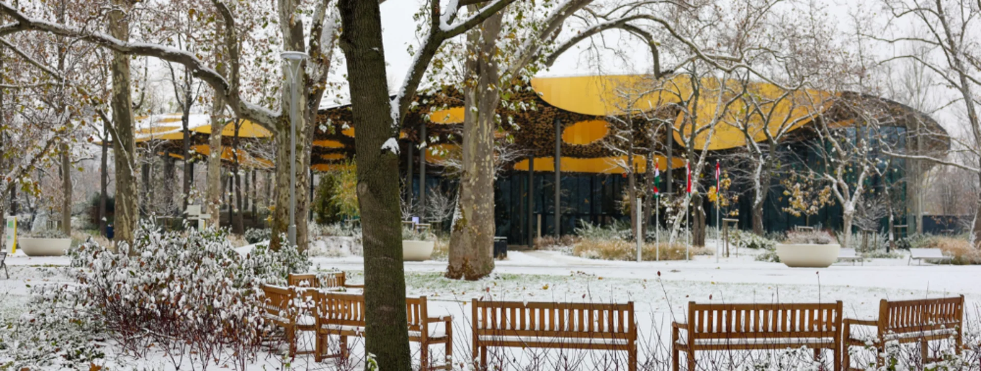 Snowy park scene with wooden benches and the House of Music, a yellow-roofed building, in the background.
