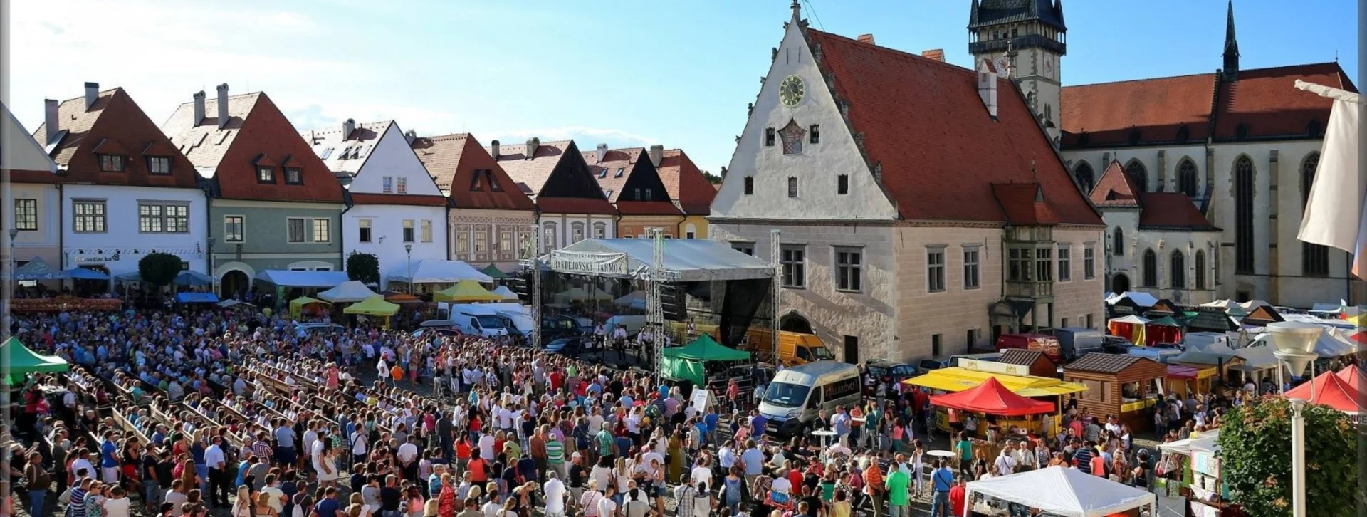 Crowded outdoor market in a historical town square with colorful stalls and traditional buildings, during the Bardejov fair.