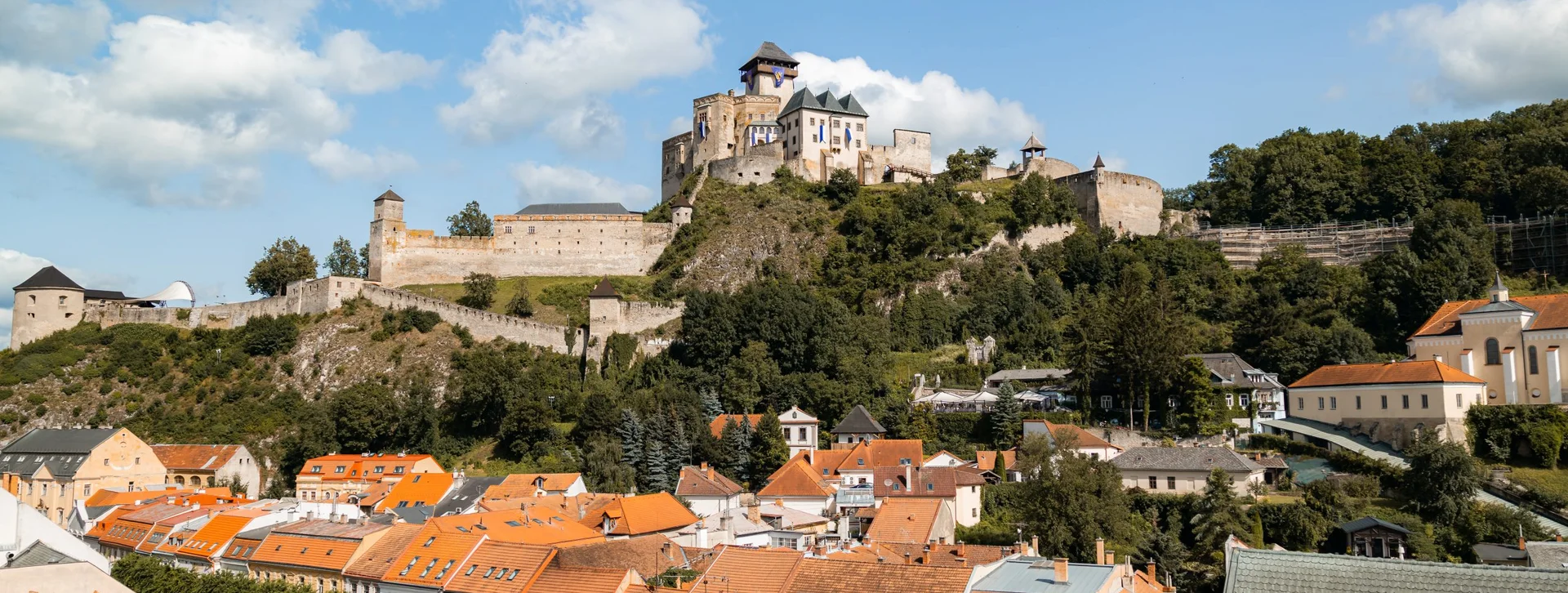 View of Trenčín Castle perched on a hill, overlooking its town with orange rooftops and surrounded by greenery under a blue sky.
