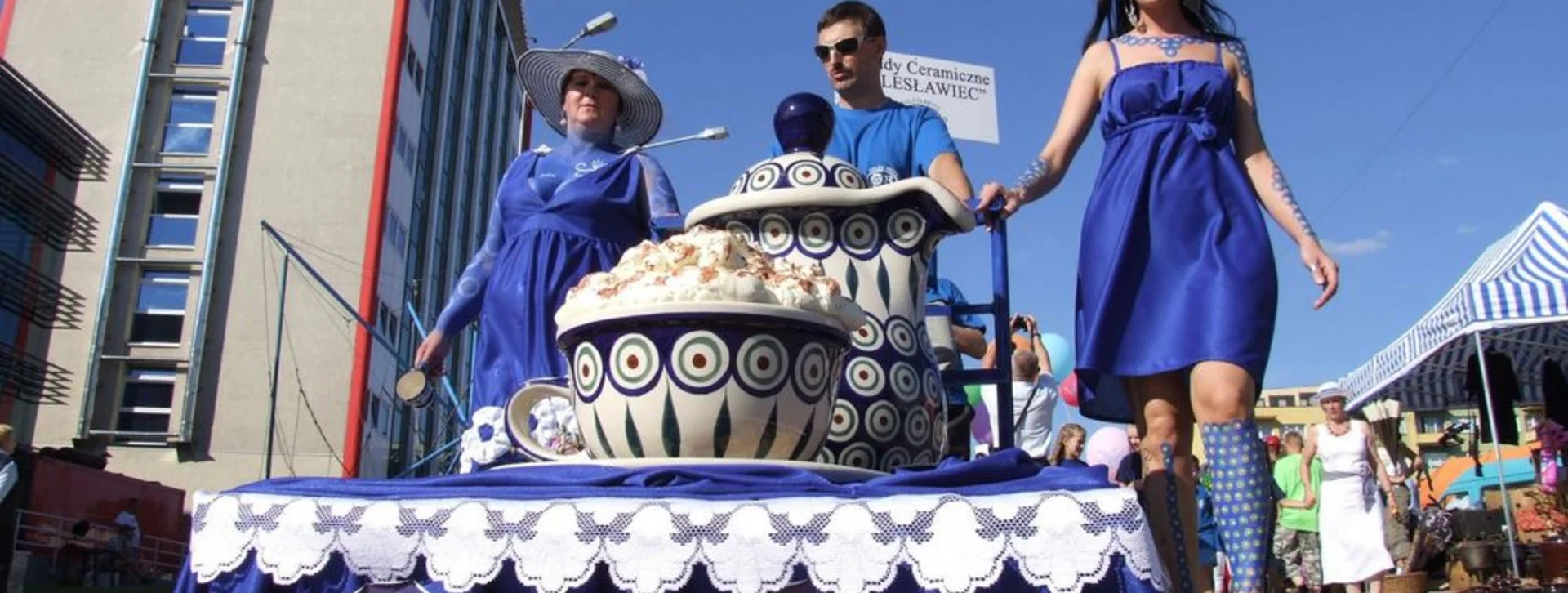 A parade scene featuring individuals in blue dresses, featuring a decorated cart with traditional pottery and food, under a sunny sky.