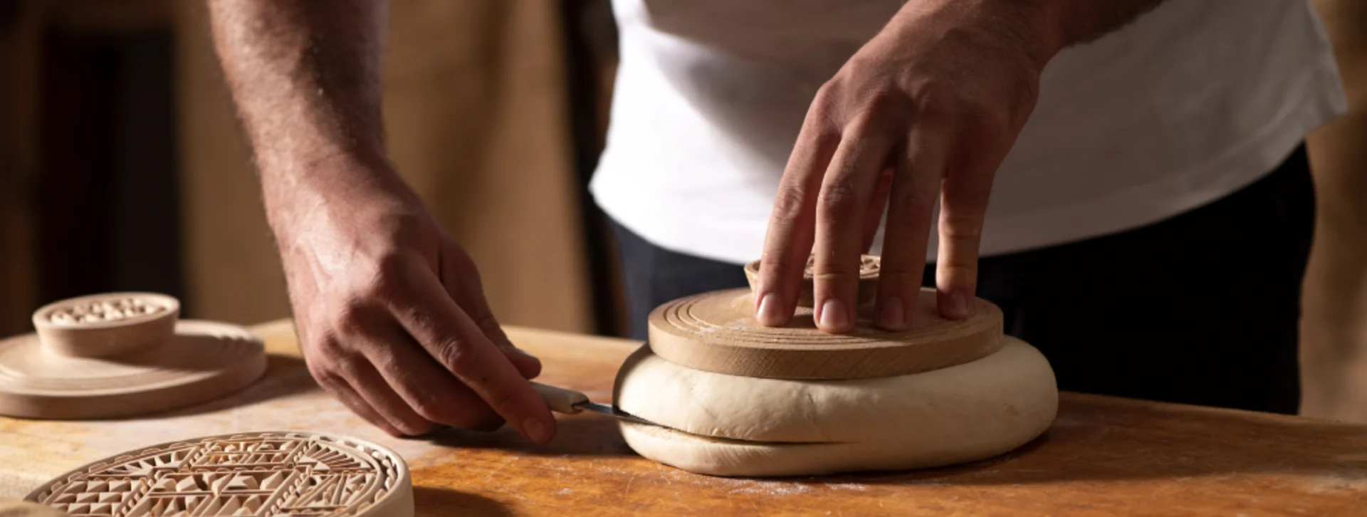A baker shapes dough on a wooden surface, pressing a patterned mold into it with one hand, while holding a knife in the other.