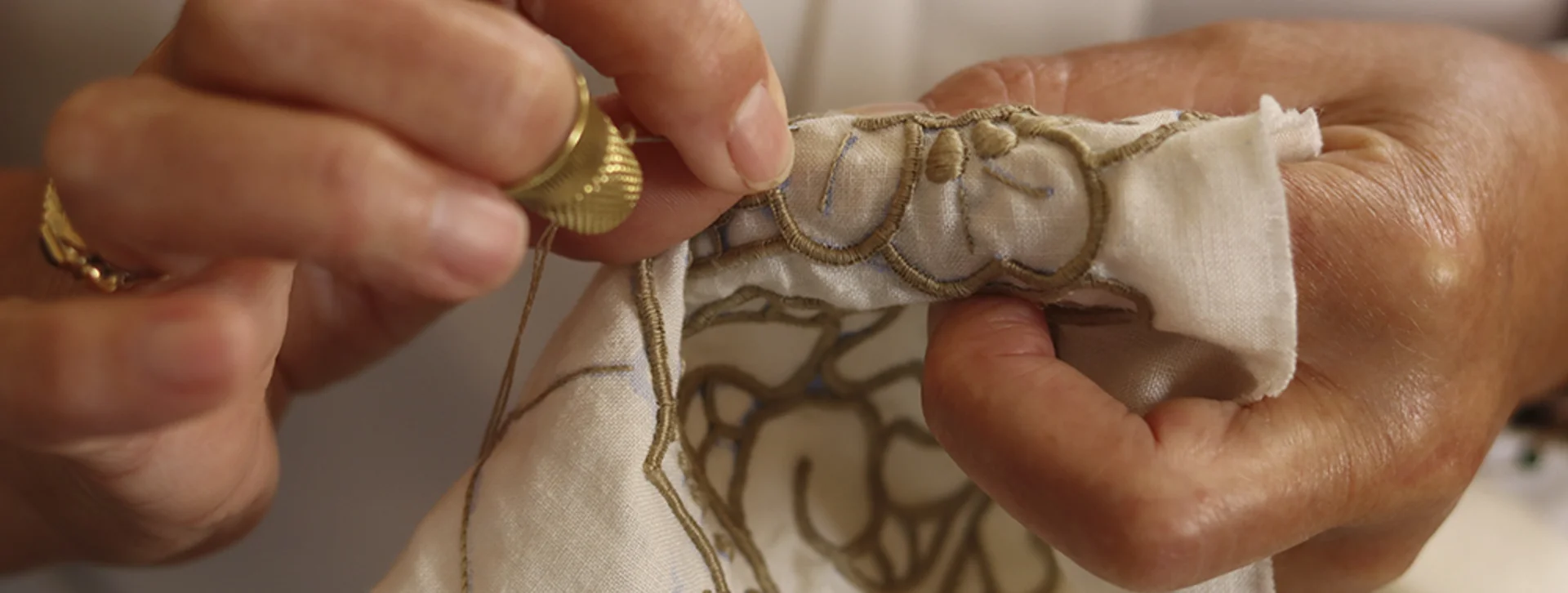 A close-up of hands sewing intricate embroidery on fabric, showcasing detailed craftsmanship and a gold thimble on the thumb.