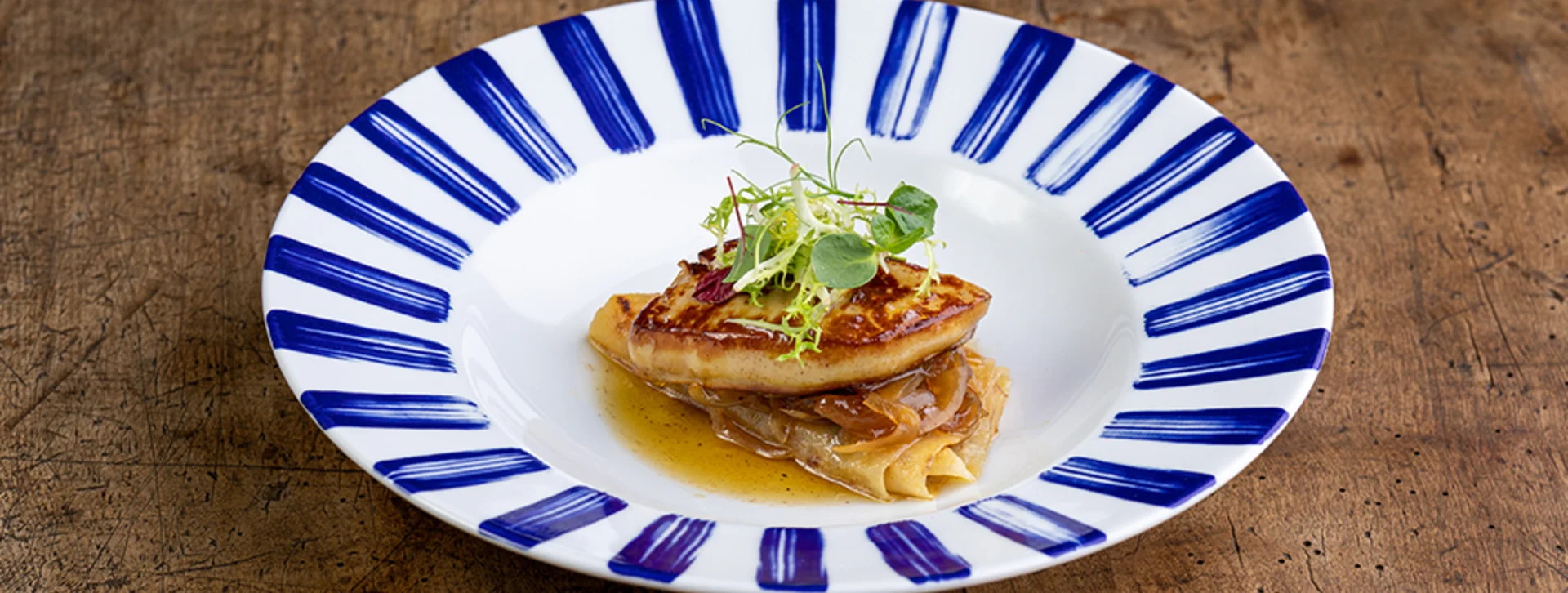 A beautifully plated dish featuring seared fish atop delicate pasta, garnished with microgreens, served on a decorative blue-striped plate.