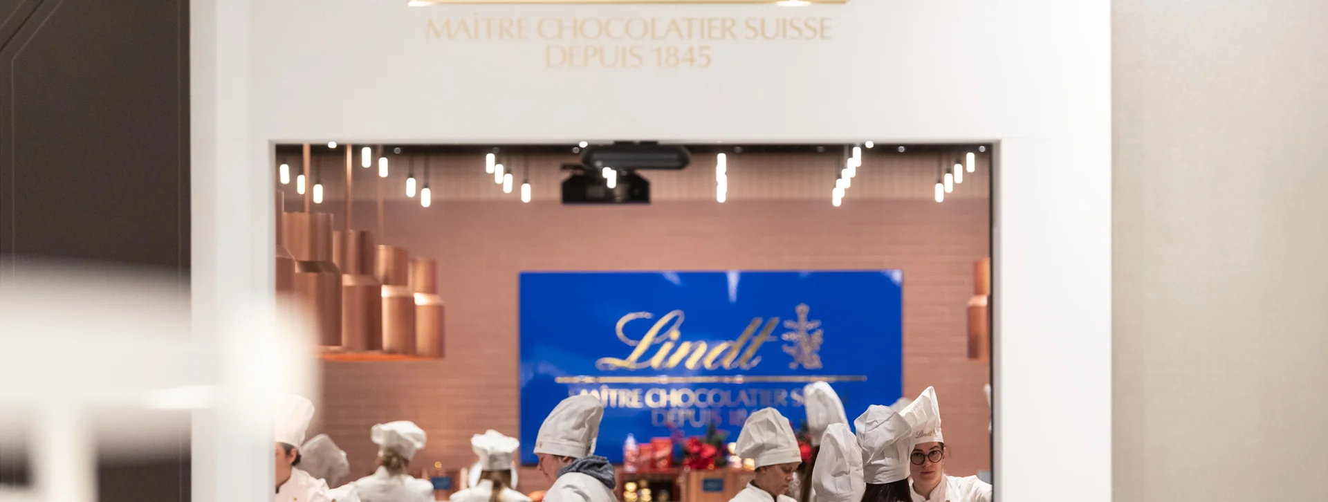 Lindt chocolate shop interior featuring chefs in white uniforms and hats working around copper pots, with a blue backdrop displaying the brand logo.