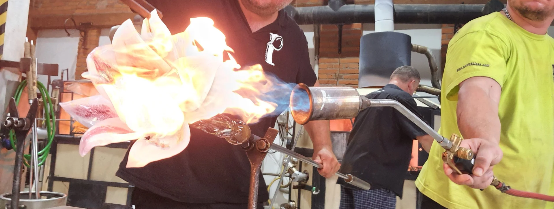Two artisans work with glass, manipulating molten material with a flame torch in a vibrant glassblowing studio.