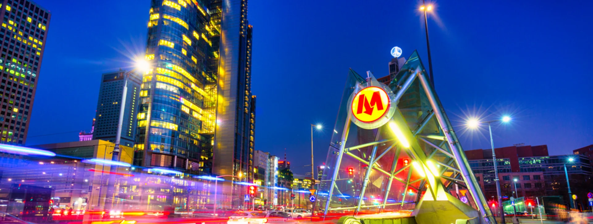 A modern metro station entrance in Warsaw illuminated at night, with skyscrapers and city traffic blurred in motion.