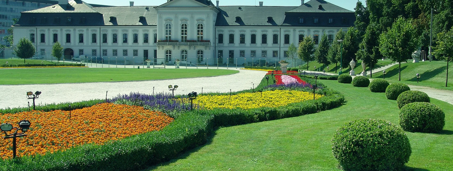 A grand white palace with a manicured lawn and vibrant flowerbeds in the foreground, set against a clear blue sky.