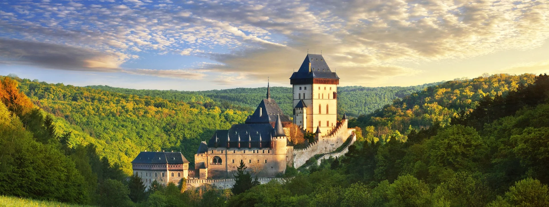 A majestic castle nestled in lush green hills under a vibrant sky filled with clouds, illuminated by warm golden sunlight.