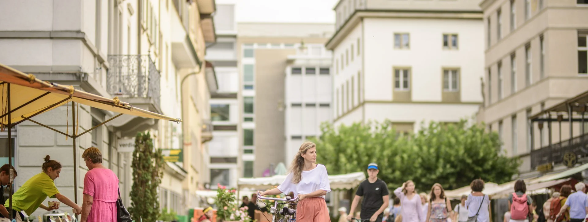 A lively market street filled with people, vibrant stalls, plants, and a woman walking with a bicycle in a pastel skirt.