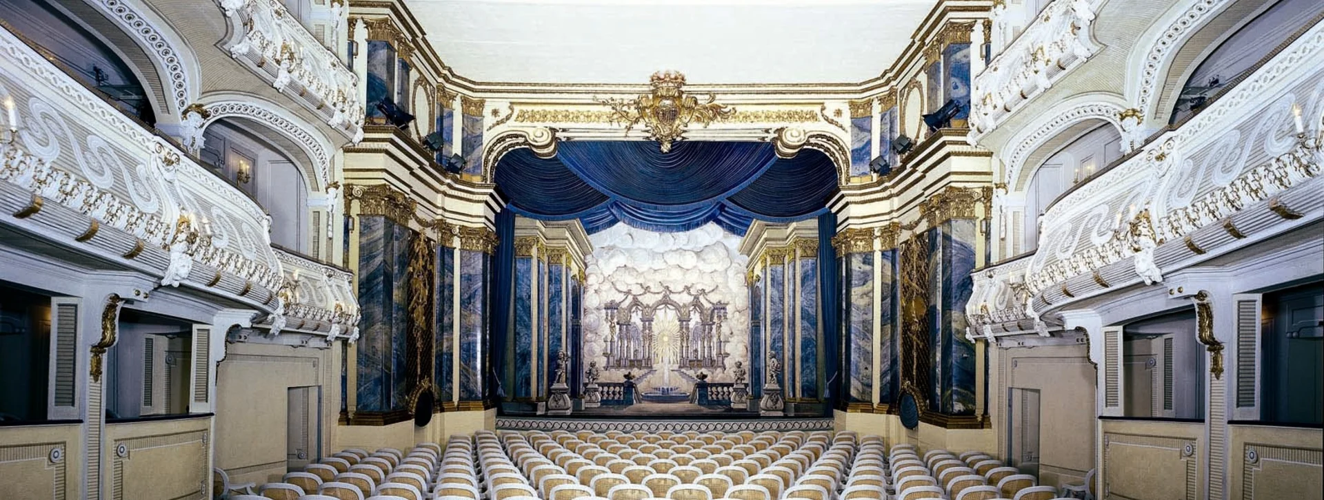 An elegant theater interior featuring rows of white cane chairs, ornate detailing, and a grand stage with blue drapery.
