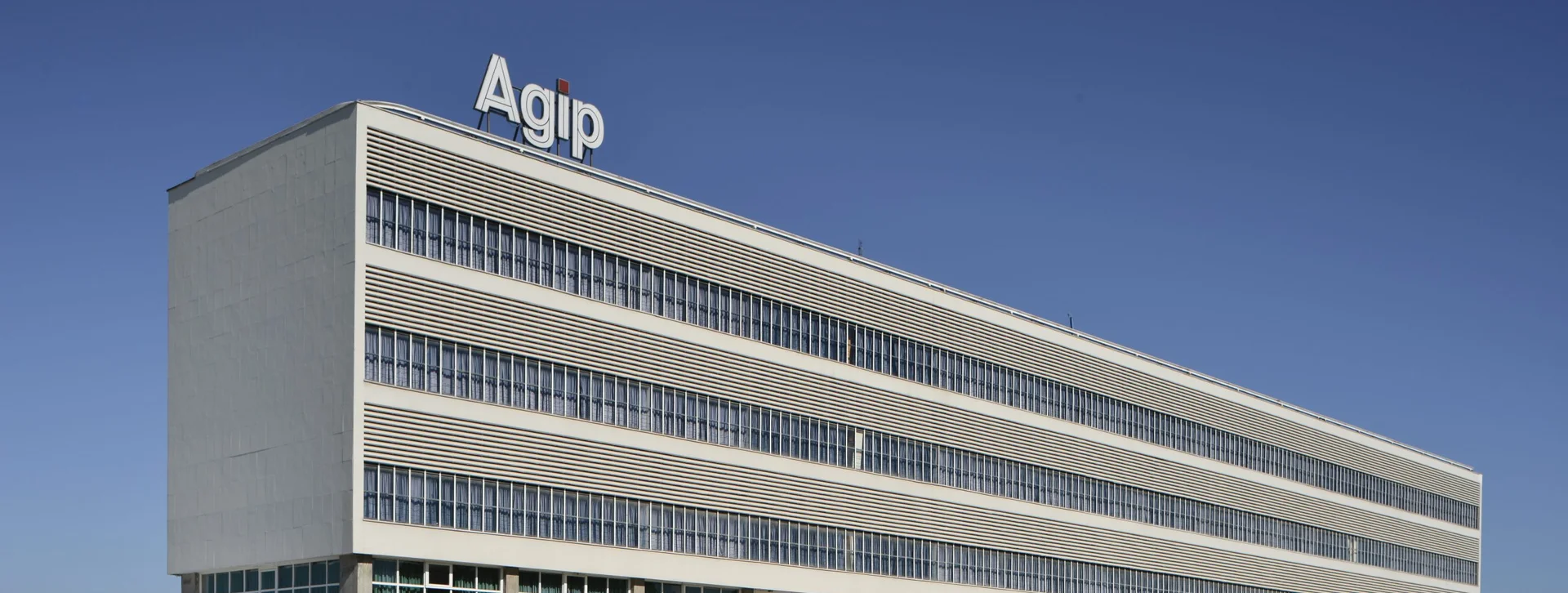 Modernist building with the Agip logo on top, featuring long horizontal windows and a clear blue sky.