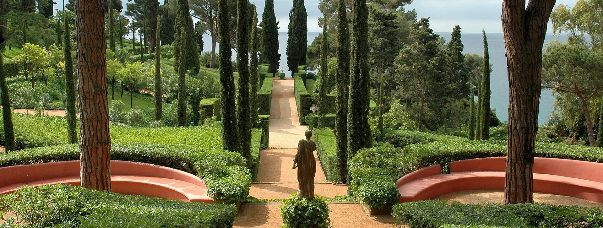 A serene garden with neatly trimmed hedges, tall cypress trees, and a statue at the center, overlooking a distant water view.