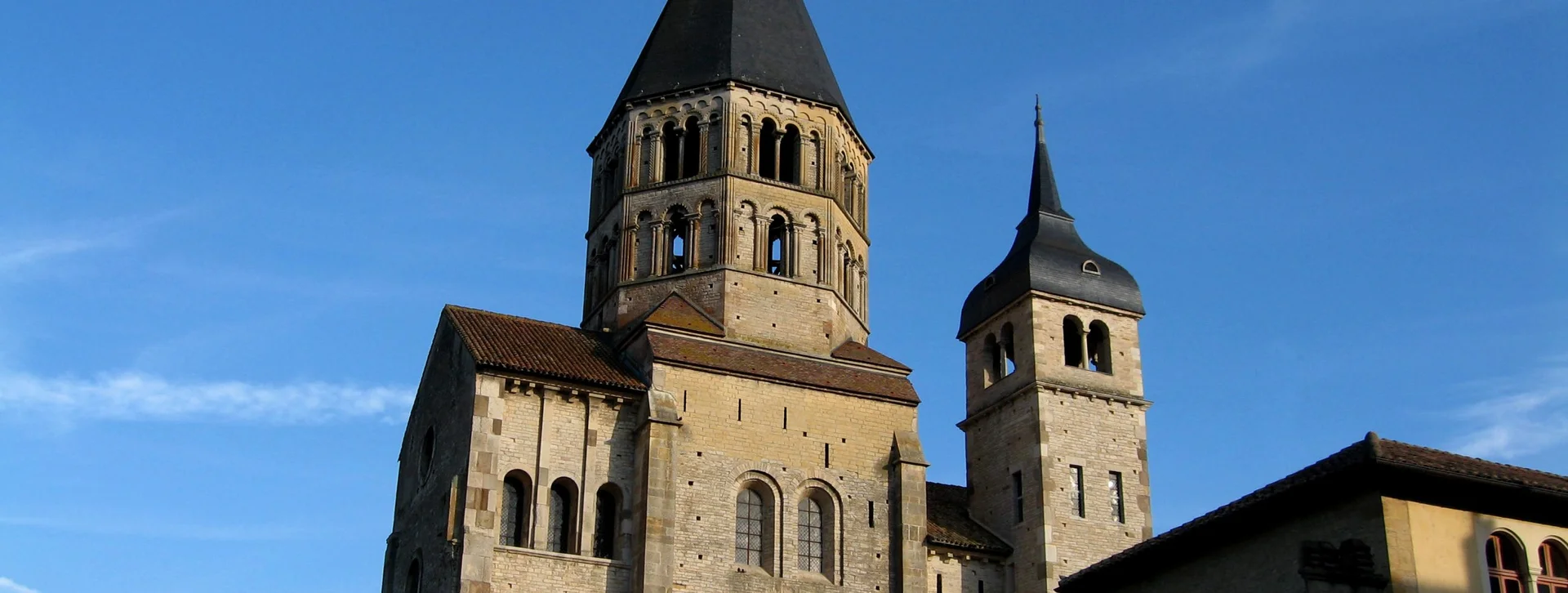 A historic stone church with two distinct towers under a clear blue sky, featuring intricate windows and a prominent cross atop the main spire.