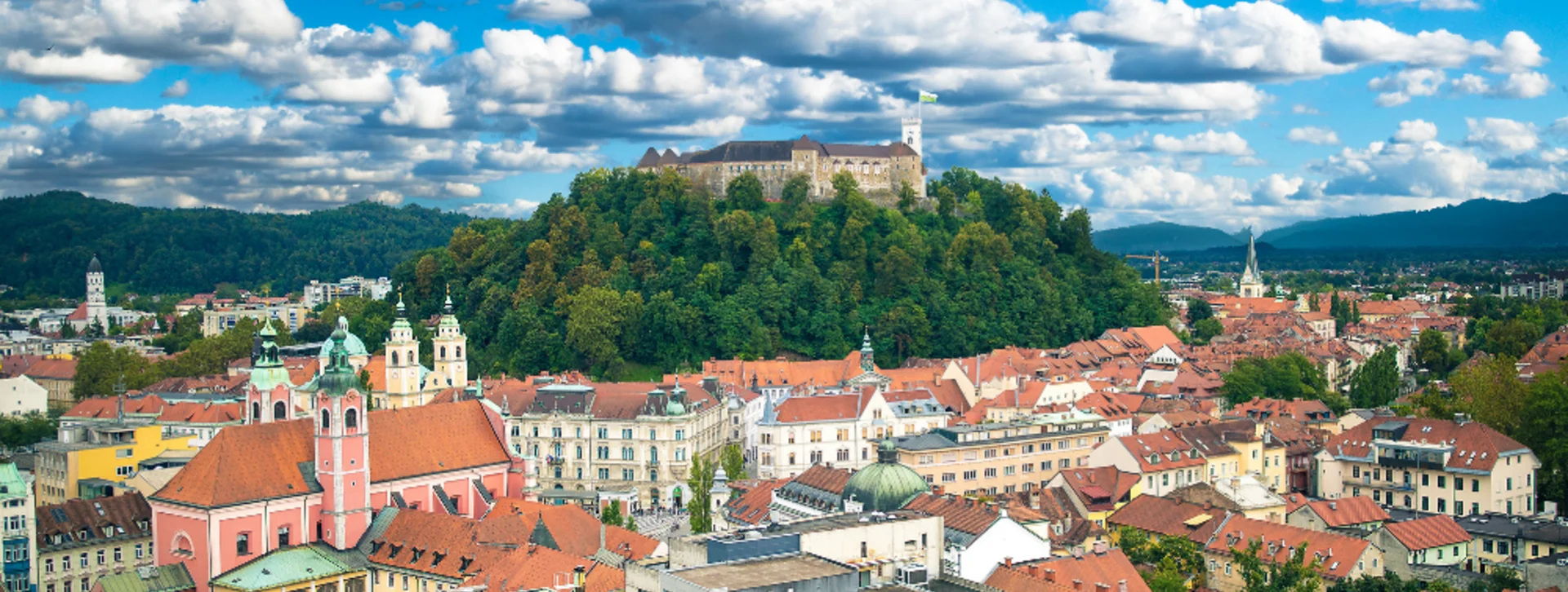 A panoramic view of Ljubljana, Slovenia, featuring the castle atop a green hill and charming red-roofed buildings under a blue sky.