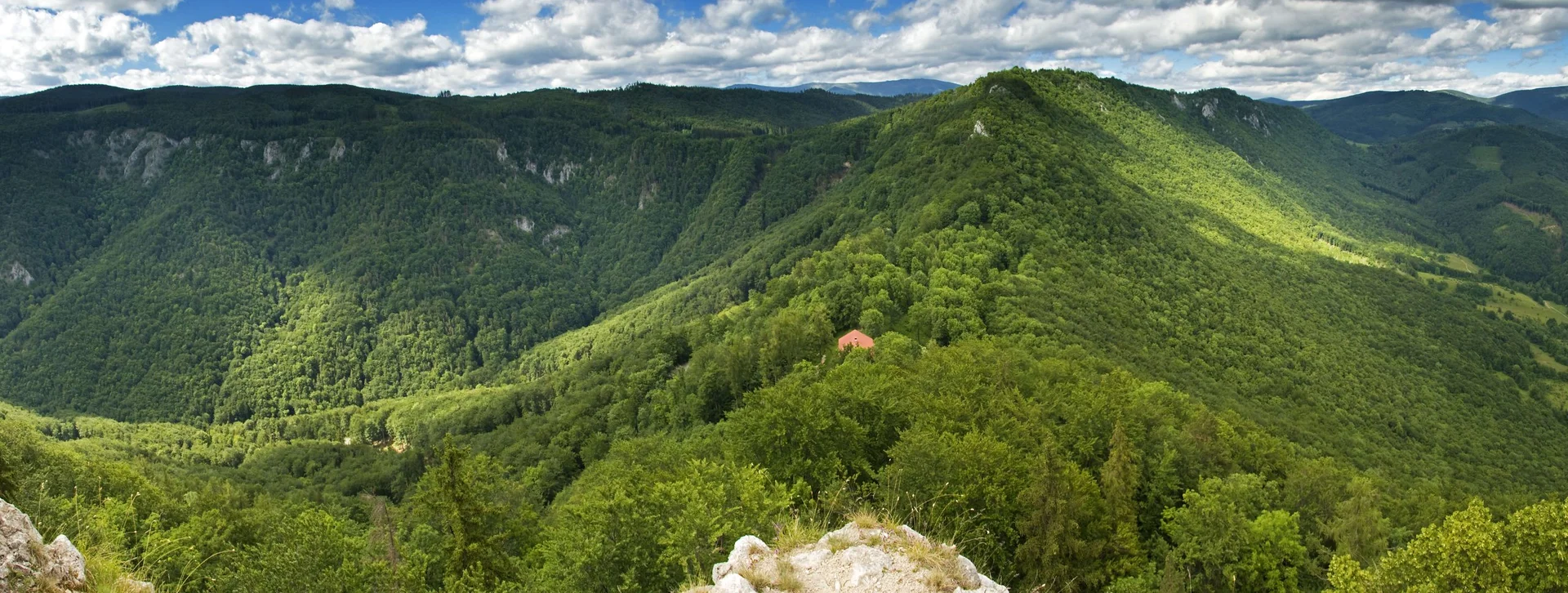 A panoramic view of lush green mountains under a partly cloudy sky, with a small red-roofed house nestled among the trees.