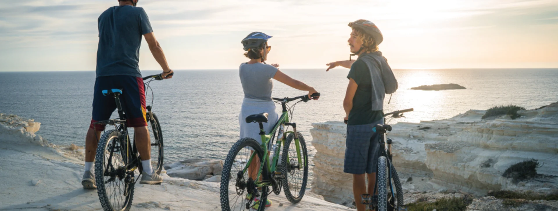 Three cyclists admire a stunning sunset over the ocean from a coastal cliff, surrounded by rugged white rock formations.