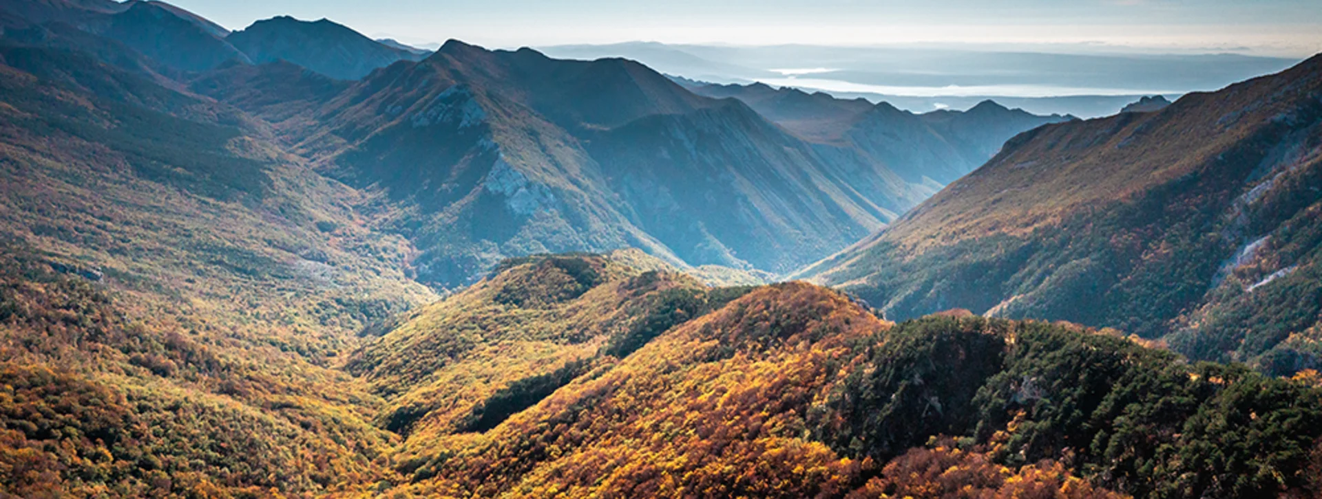 Vast mountain landscape with rolling hills covered in autumn foliage under a clear blue sky, extending into the distant horizon.